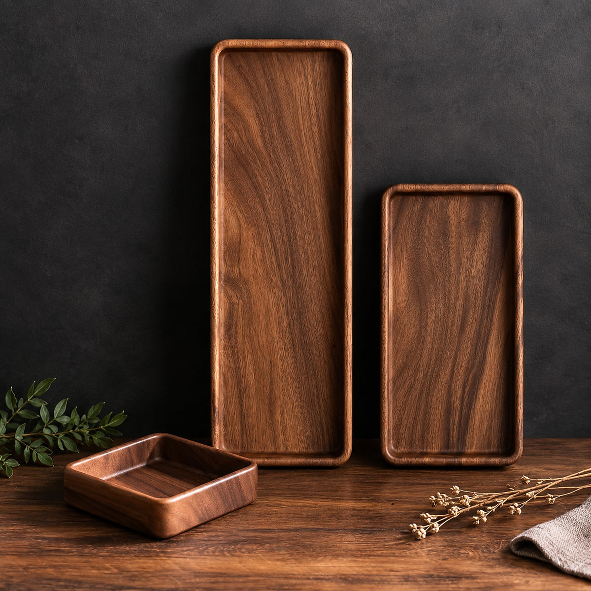 Three rectangular wooden serving trays with rounded corners in two heights and one small square tray, arranged on a wooden surface against a dark textured wall with dried flowers and greenery as props.