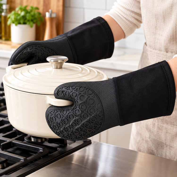 A person wearing black silicone oven mitts with textured grip panels holds a cream-colored Dutch oven on a stovetop in a bright kitchen setting.