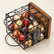 A black metal wire coffee capsule organizer with a wooden back panel, filled with assorted coffee capsules in gold, bronze, red, and brown tones, on a light beige background.