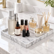 A marble bathroom vanity tray holding assorted skincare and beauty products, including bottles, tubes, lipsticks, a perfume bottle, and a reed diffuser, arranged on a white marble countertop with gold-toned fixtures in the background.