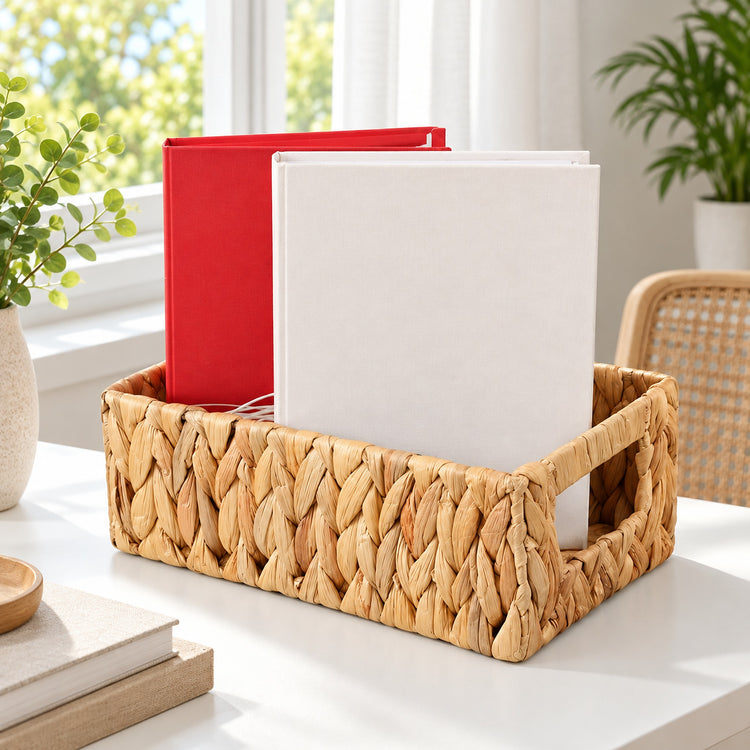 A woven natural-fiber desk organizer tray holding two binders, one red and one white, on a white tabletop in a bright room with plants and a chair visible.