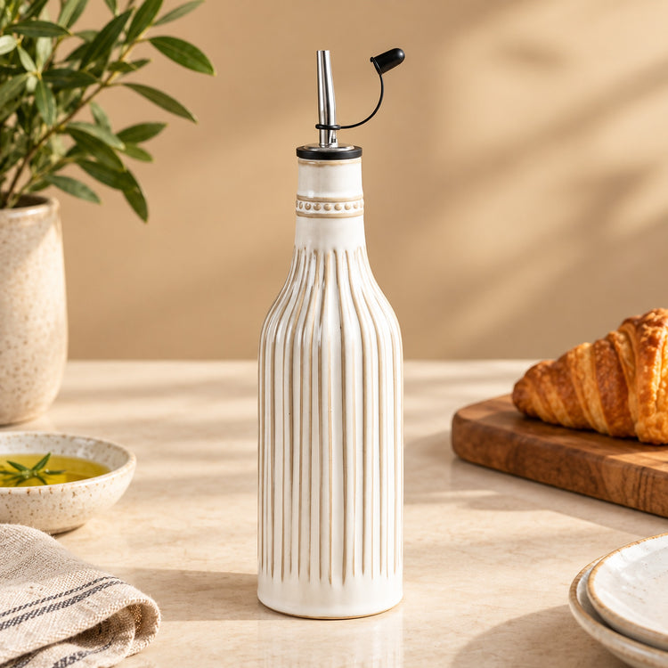 A ribbed ceramic oil bottle with a silver metal pour spout and black flip-top lid, styled on a beige tabletop with a croissant, bowl of oil, and greenery in the background.