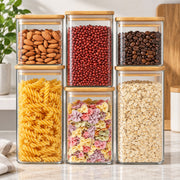 Six clear glass canisters with light wood lids are arranged in two rows on a countertop, filled with almonds, red beans, coffee beans, rotini pasta, multicolor bowtie pasta, and oats. A plant and kitchen background are visible.