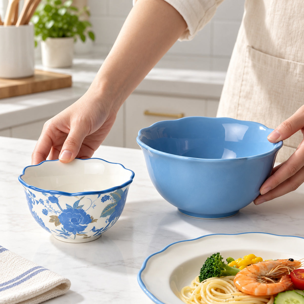 A person in a beige apron holds two ceramic bowls on a white marble kitchen counter: a large light blue bowl and a smaller white bowl with blue floral patterns and a blue rim. A plate of pasta, shrimp, broccoli, and tomato is partly visible in the foreground.