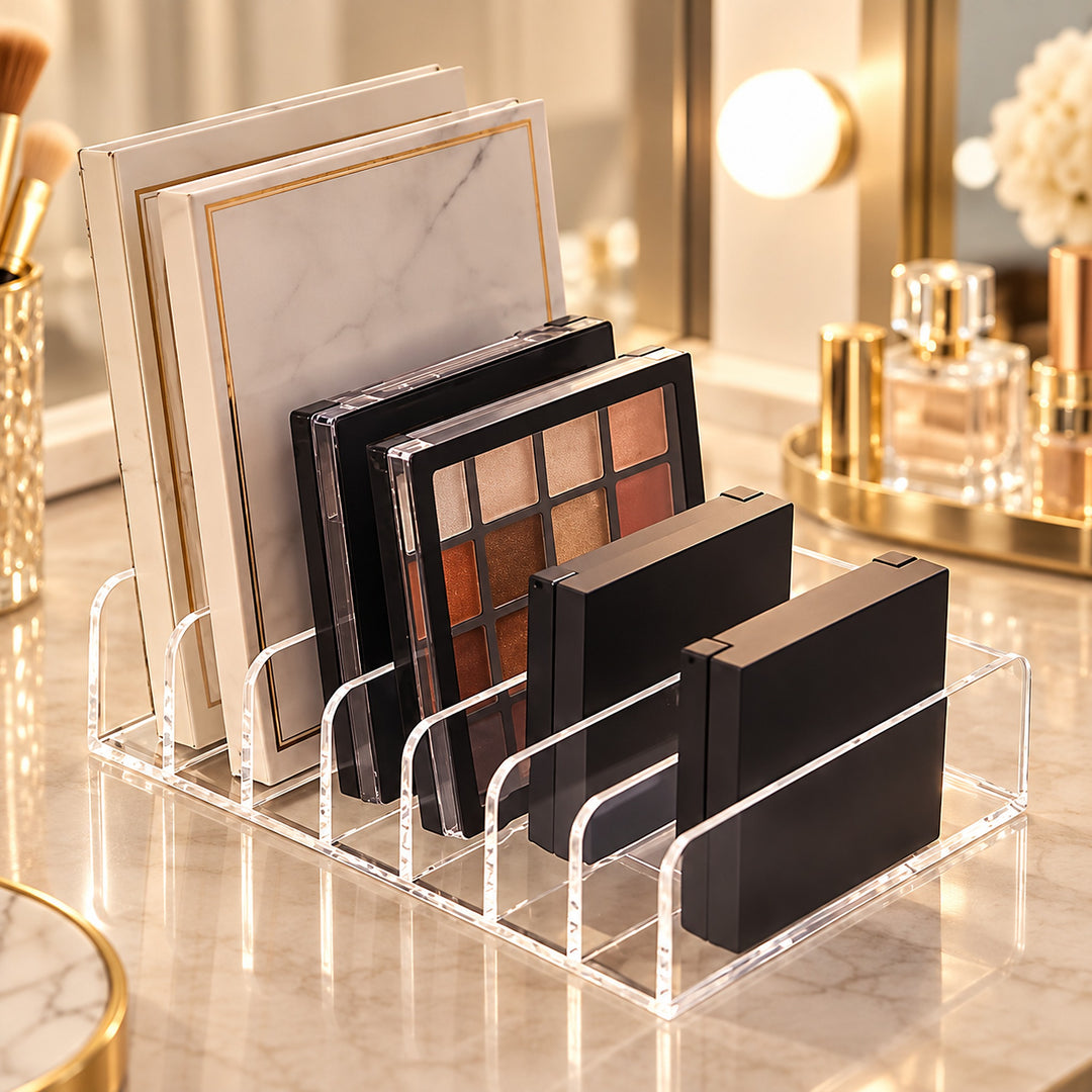 Clear acrylic makeup organizer stand holding several eyeshadow palettes and compact cases on a marble vanity surface in a softly lit bathroom or makeup station setting.