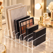 Clear acrylic makeup organizer stand holding several eyeshadow palettes and compact cases on a marble vanity surface in a softly lit bathroom or makeup station setting.