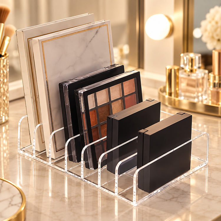 Clear acrylic makeup organizer stand holding several eyeshadow palettes and compact cases on a marble vanity surface in a softly lit bathroom or makeup station setting.