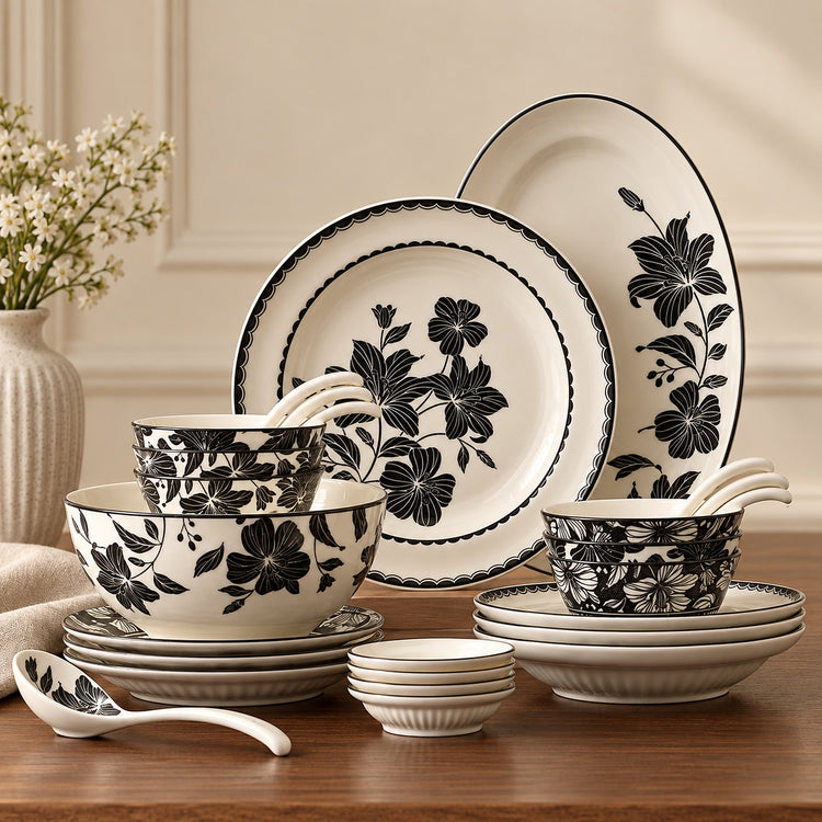 A coordinated ceramic dinnerware set with black floral and scalloped border designs on an off-white background, arranged on a wooden table with a vase of flowers in the background.