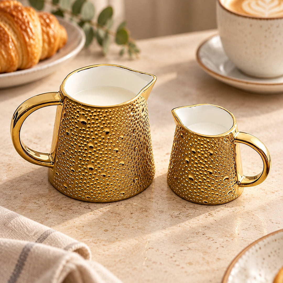 A gold ceramic milk pitcher and matching creamer set with a textured, dimpled exterior and smooth white interiors, styled on a beige countertop with a croissant, coffee cup, and cloth napkin in the background.