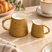 A gold ceramic milk pitcher and matching creamer set with a textured, dimpled exterior and smooth white interiors, styled on a beige countertop with a croissant, coffee cup, and cloth napkin in the background.