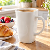A tall white ceramic mug filled with coffee sits on a wooden table in a bright kitchen setting, with a croissant on a plate and fresh berries nearby.