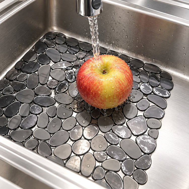 A red and yellow apple sits in a stainless steel kitchen sink with a faucet running over it. The sink floor is covered with dark gray pebble-style drain covers.