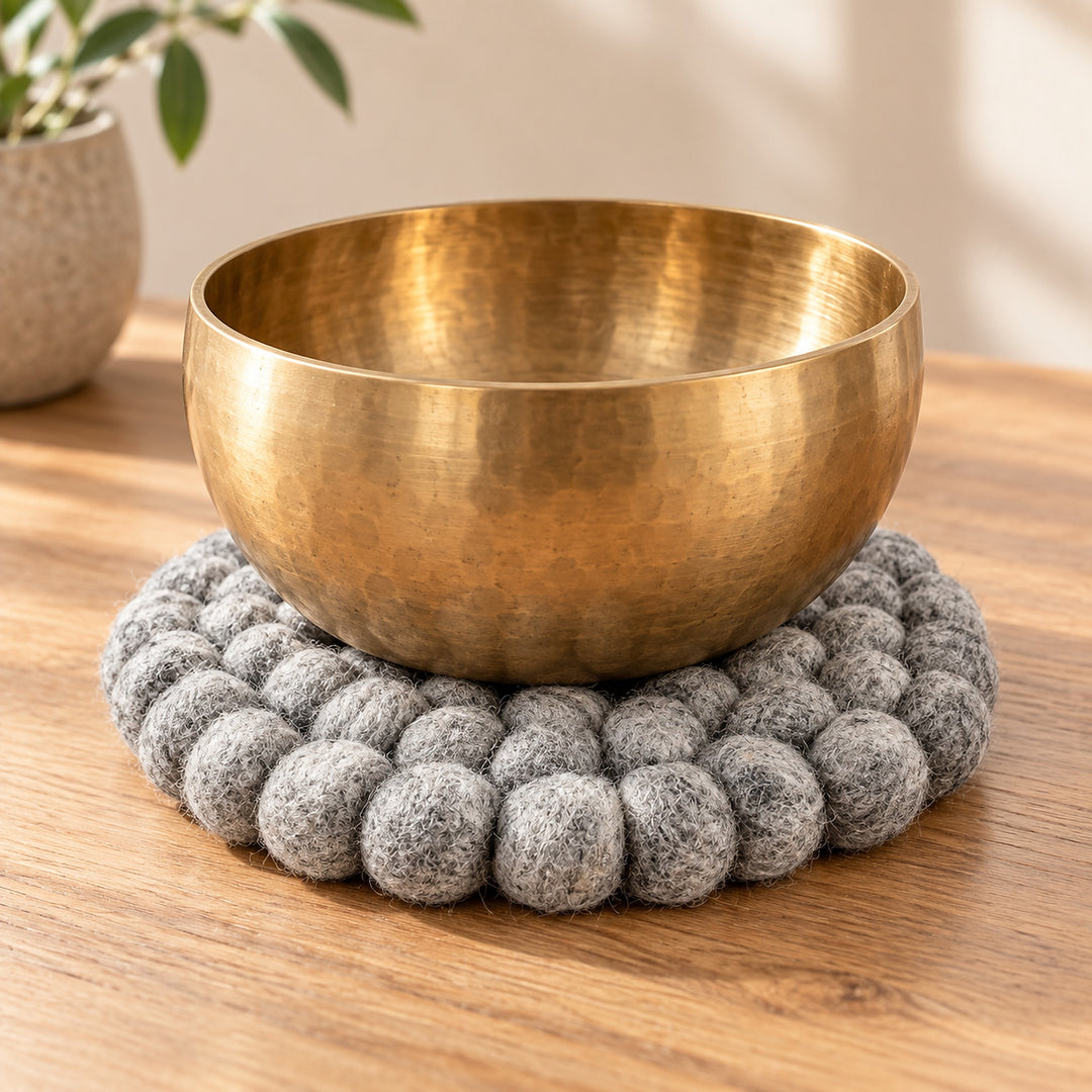 A hammered brass singing bowl resting on a round gray felt coaster on a wooden table, with a blurred potted plant in the background.