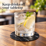 A clear glass tumbler with a thick base sits on a round black marble coaster on a wooden table. The glass contains a clear bubbly drink with ice, a lemon slice, and mint leaves.