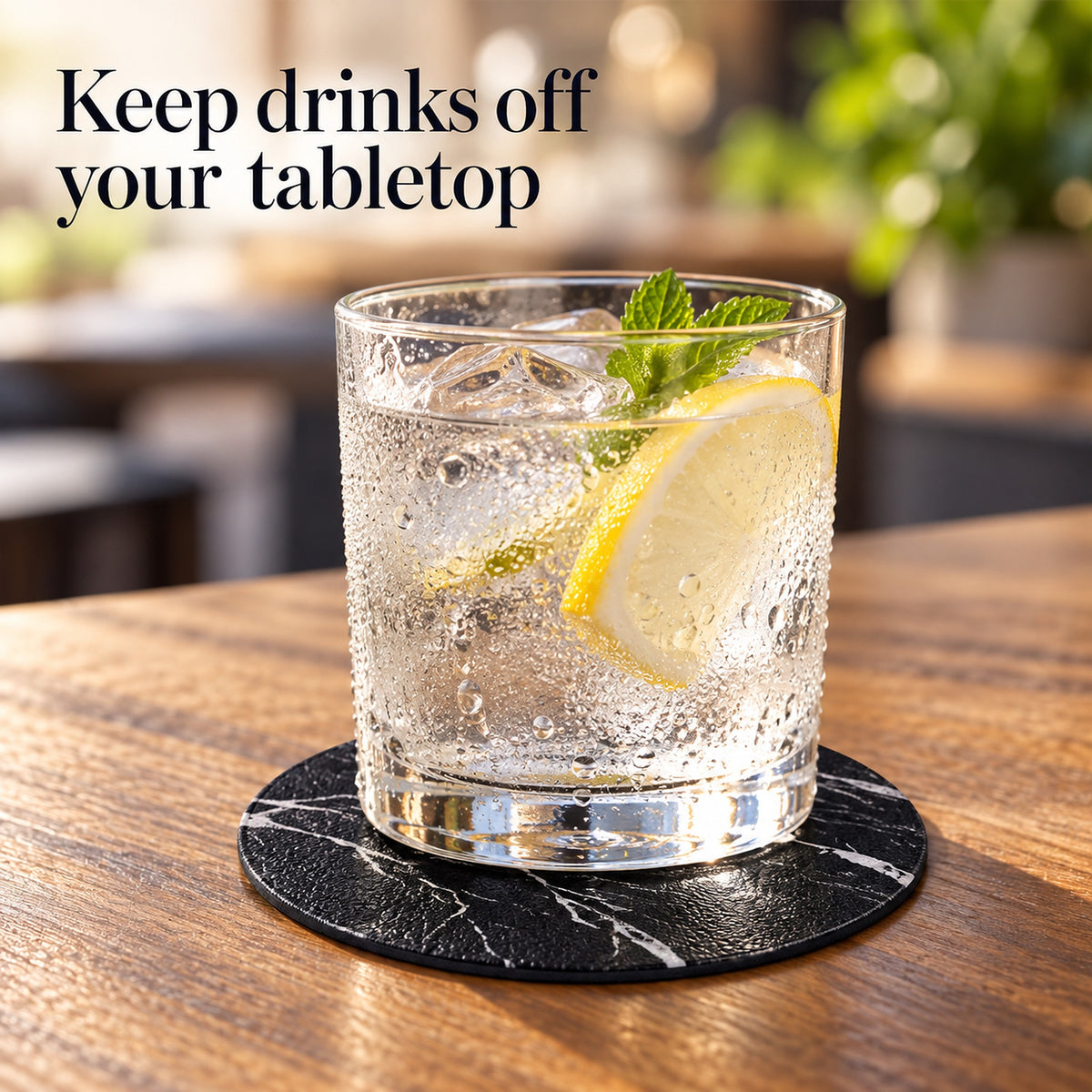 A clear glass tumbler with a thick base sits on a round black marble coaster on a wooden table. The glass contains a clear bubbly drink with ice, a lemon slice, and mint leaves.