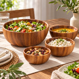 A set of five wooden bowls of varying sizes arranged on a dining table, filled with a large salad, soup, rice, and nuts, set against a warm, softly blurred home dining background.