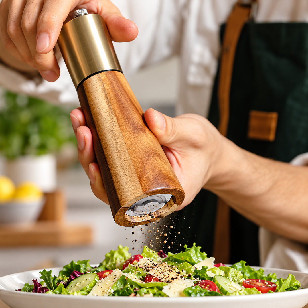 Acacia wood and gold-colored pepper grinder used over a fresh salad.
