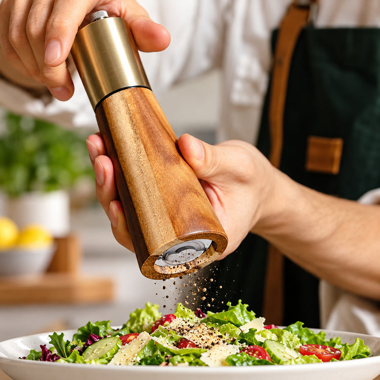 Acacia wood and gold-colored pepper grinder used over a fresh salad.