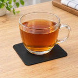 Black square silicone coaster under a glass cup of tea on a wooden table.