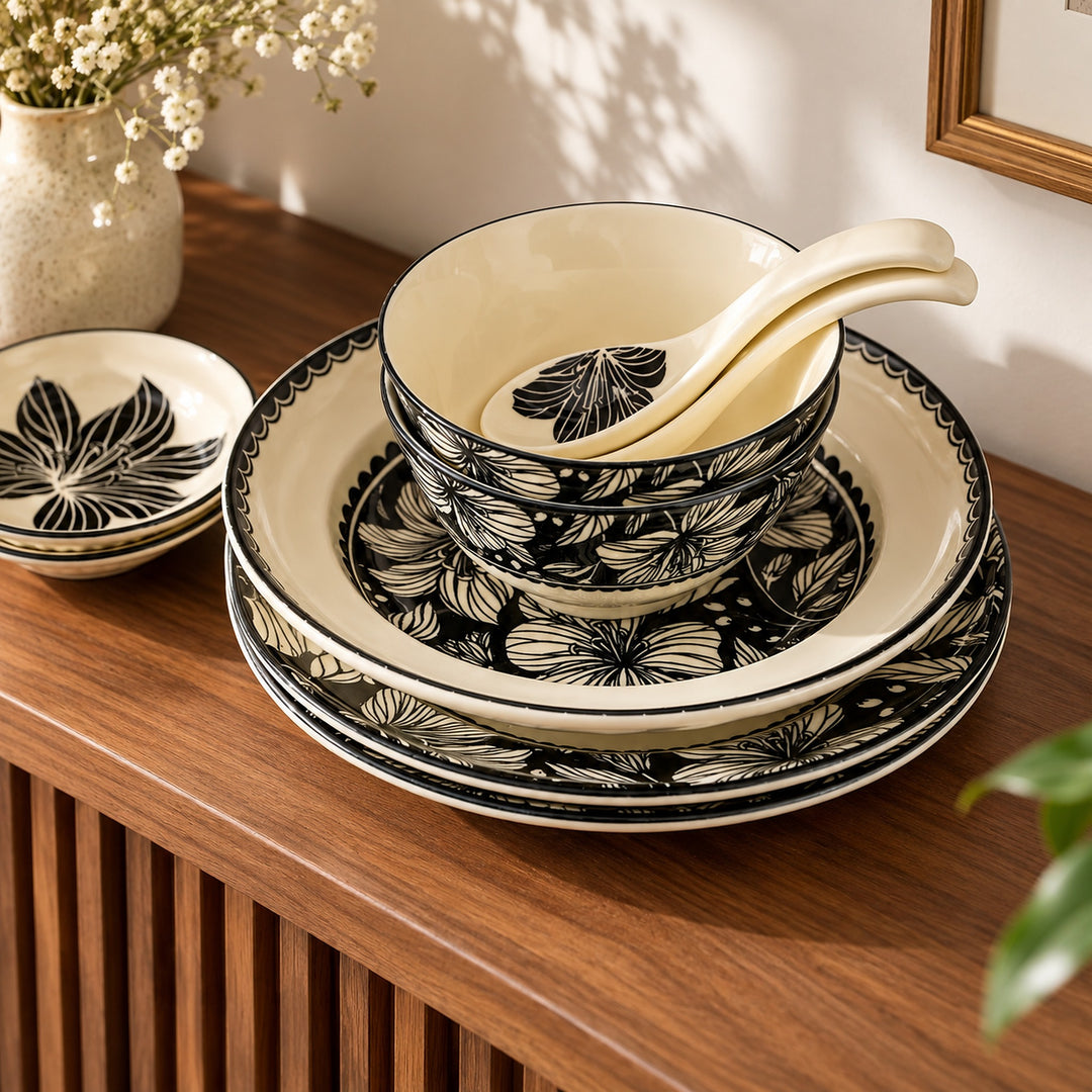 A ceramic dinnerware set with a black and cream floral pattern, including plates, bowls, and a soup spoon, arranged on a wooden table next to a vase of small white flowers.