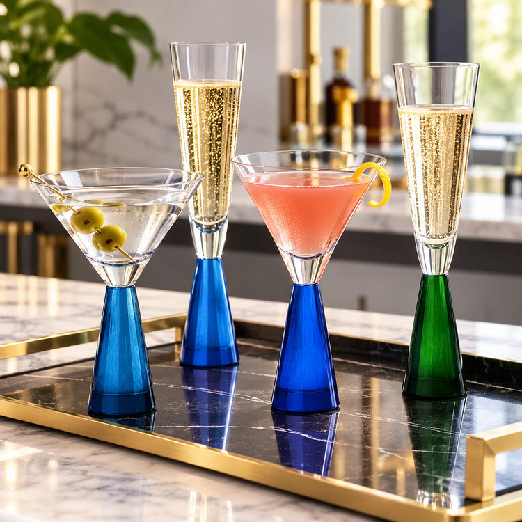 Four elegant cocktail glasses with colorful glass bases arranged on a black marble tray with gold trim in a bar setting. The glasses include a martini glass with olives, a tall champagne flute with bubbly golden liquid, a pink cocktail flute, and a green champagne flute.