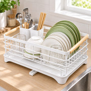 A white dish drying rack with light wood handles sits on a kitchen counter, holding stacked white and green plates, bowls, and clear drinking glasses. A sink and window are visible in the background.