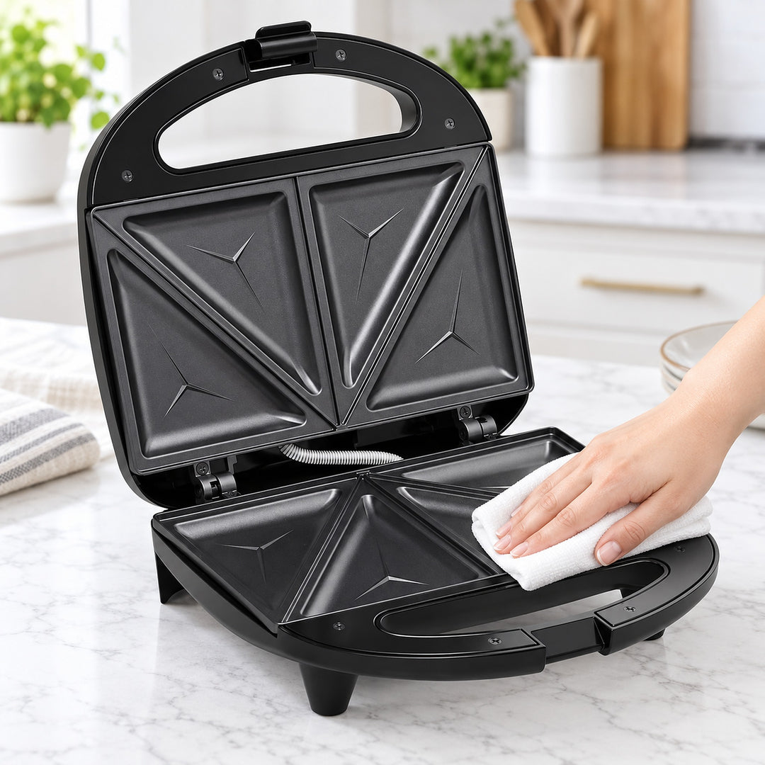 A black electric sandwich maker/triple-decker toaster is open on a white marble kitchen countertop, with a person's hand wiping the inside with a white cloth. The background shows a blurred modern kitchen setting.