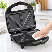 A black electric sandwich maker/triple-decker toaster is open on a white marble kitchen countertop, with a person's hand wiping the inside with a white cloth. The background shows a blurred modern kitchen setting.
