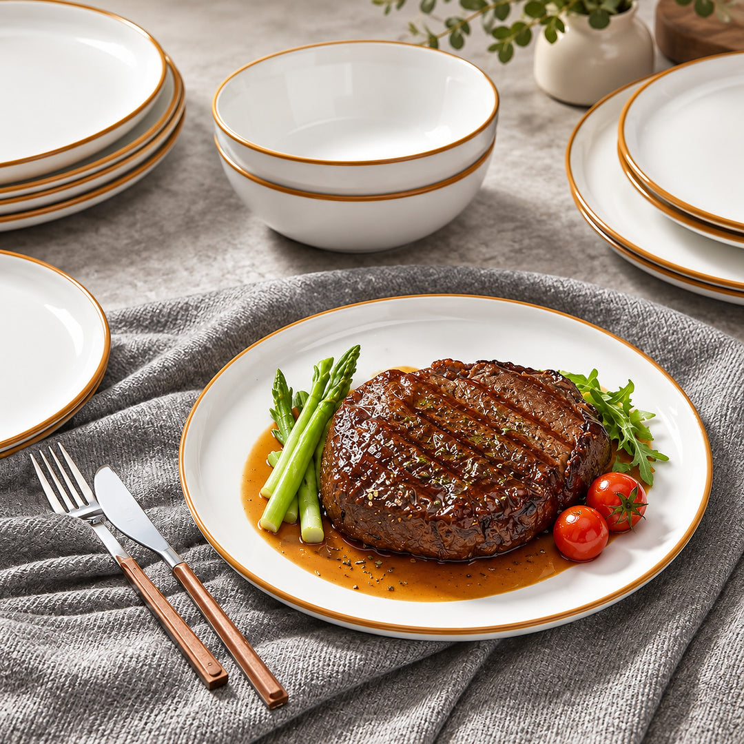 A white ceramic dinner plate with a gold rim, styled with a grilled steak, asparagus, cherry tomatoes, and a knife on a gray textured tablecloth.