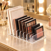 Clear acrylic makeup organizer stand holding multiple eyeshadow palettes and a blush compact on a vanity counter, with a mirror and makeup brushes in the background.