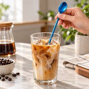 A hand holds a bright blue spoon over a clear glass of iced coffee with milk swirls on a marble countertop, with coffee beans, a glass bottle of coffee, and a folded cloth nearby.