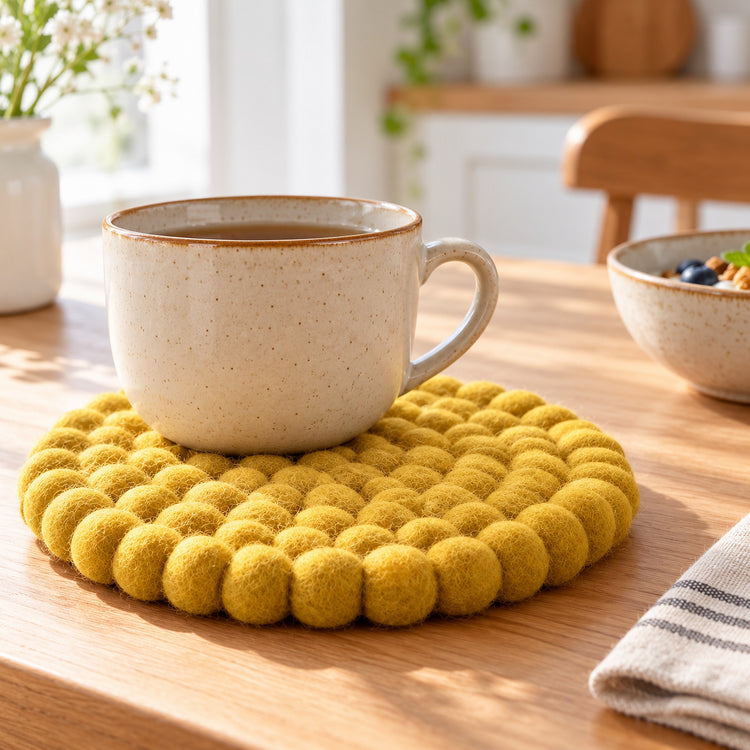 A round yellow felt pom-pom trivet coaster sits on a wooden table under a speckled beige ceramic mug, with a softly blurred kitchen background.