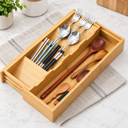 A bamboo utensil organizer tray with multiple compartments holding silver forks, spoons, chopsticks, and wooden spoons, placed on a folded beige cloth over a white marble surface.