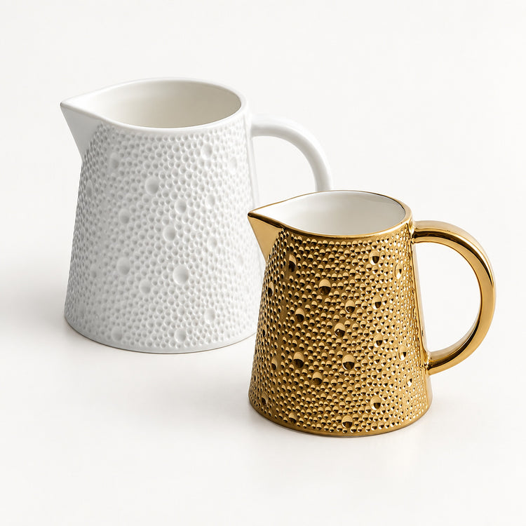 Two ceramic pitchers with a textured, dimpled surface on a clean white background. One pitcher is solid white, and the other is metallic gold with a white interior.