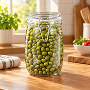 A clear glass clamp-lid jar filled with green peas, set on a wooden kitchen countertop with a blurred kitchen background.