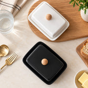 Two rectangular butter dishes with lids, one black and one white, each with a round wooden knob, arranged on a light beige tabletop with a wooden board, glass, spoon, fork, bread, and butter in a styled flat-lay setting.