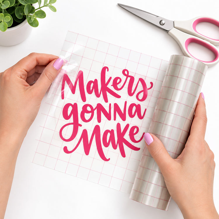 Hands holding a roll of clear vinyl or craft transfer sheet with pink grid lines and a bright pink script quote reading “Makers gonna make,” with scissors and a small potted plant on a white background.
