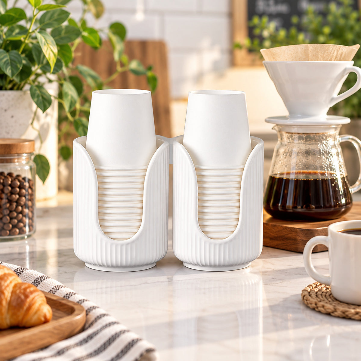 A pair of white ribbed cup holders on a kitchen counter, each holding a white cup. The scene includes a pour-over coffee maker, a coffee carafe with brewed coffee, a mug on a coaster, and plants in the background.