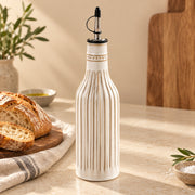 A ribbed ceramic oil bottle with a black pour spout, standing on a beige countertop in a warm kitchen setting with bread, olive oil, and a cutting board in the background.