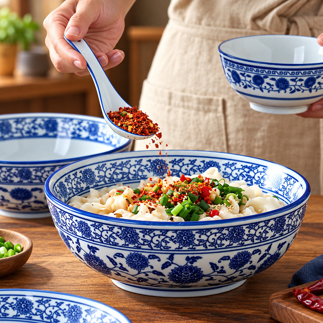 A large ceramic bowl with blue floral patterns is filled with dumplings topped with red chili flakes and green onions, while a hand holds a matching spoon above it. A smaller matching bowl is held in the other hand, and additional bowls are visible on a wooden table.