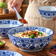 A large ceramic bowl with blue floral patterns is filled with dumplings topped with red chili flakes and green onions, while a hand holds a matching spoon above it. A smaller matching bowl is held in the other hand, and additional bowls are visible on a wooden table.