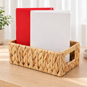 A woven natural-fiber storage basket with side handles sits on a light wood table, holding two upright files or folders, one red and one white, against a softly lit indoor background.