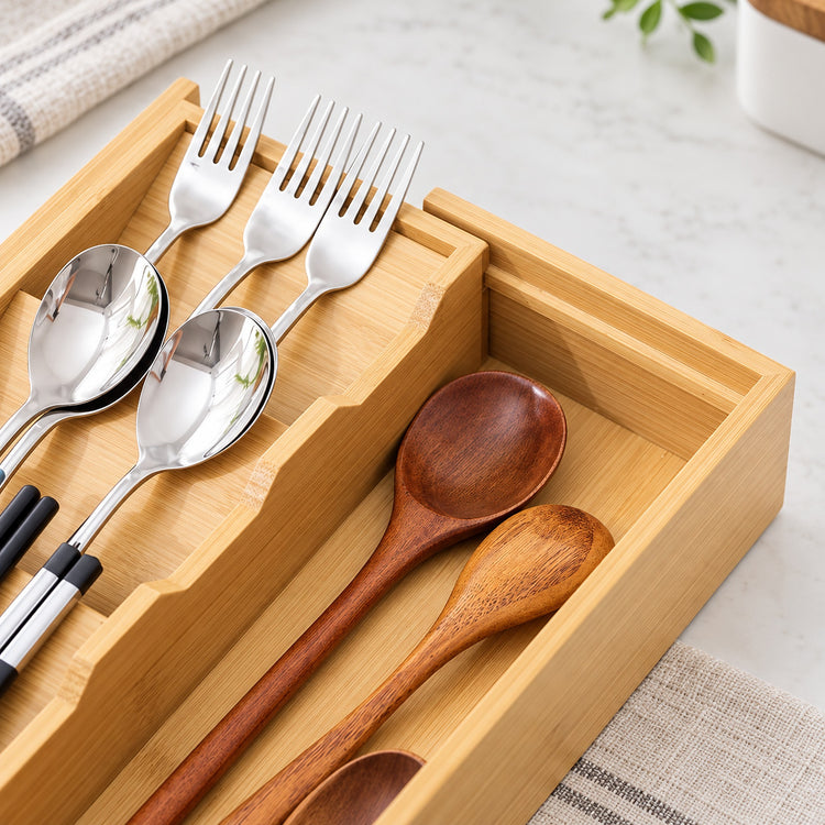A bamboo utensil organizer tray holding silver forks, spoons, chopsticks, and wooden spoons on a light marble surface.