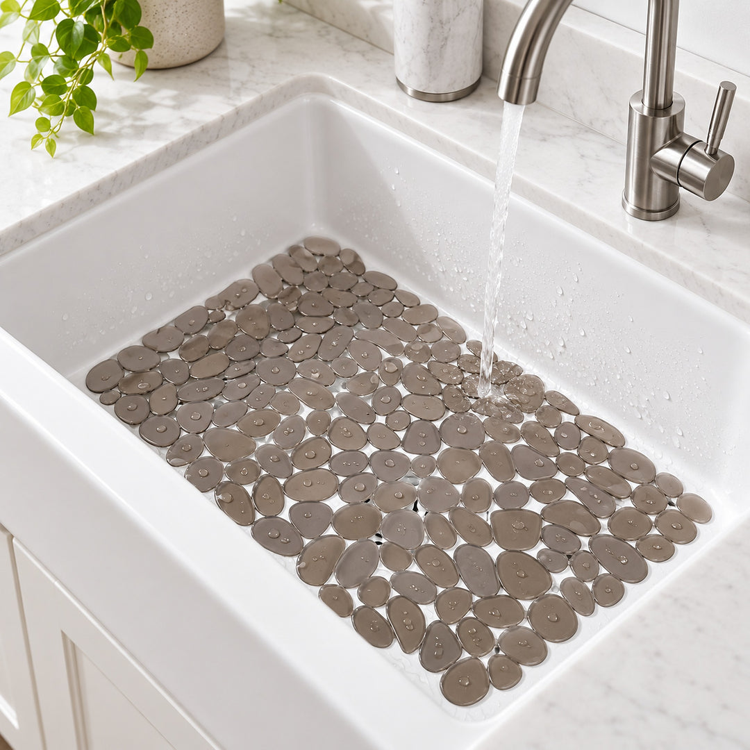 A white farmhouse-style kitchen sink with a waterfall faucet, filled with a protective mat of round gray pebble tiles. Water is running into the sink, and a marble countertop and a small green plant are visible in the background.