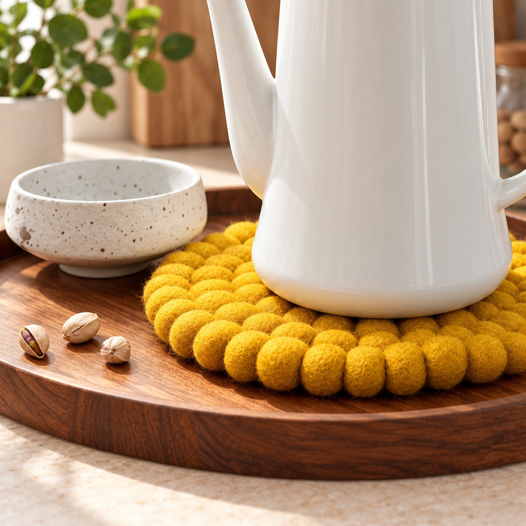 A white ceramic teapot sits on a round yellow pom-pom textured coaster on a wooden tray, next to a speckled beige ceramic cup with three pistachios on a light beige surface.