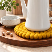 A white ceramic teapot sits on a round yellow pom-pom textured coaster on a wooden tray, next to a speckled beige ceramic cup with three pistachios on a light beige surface.