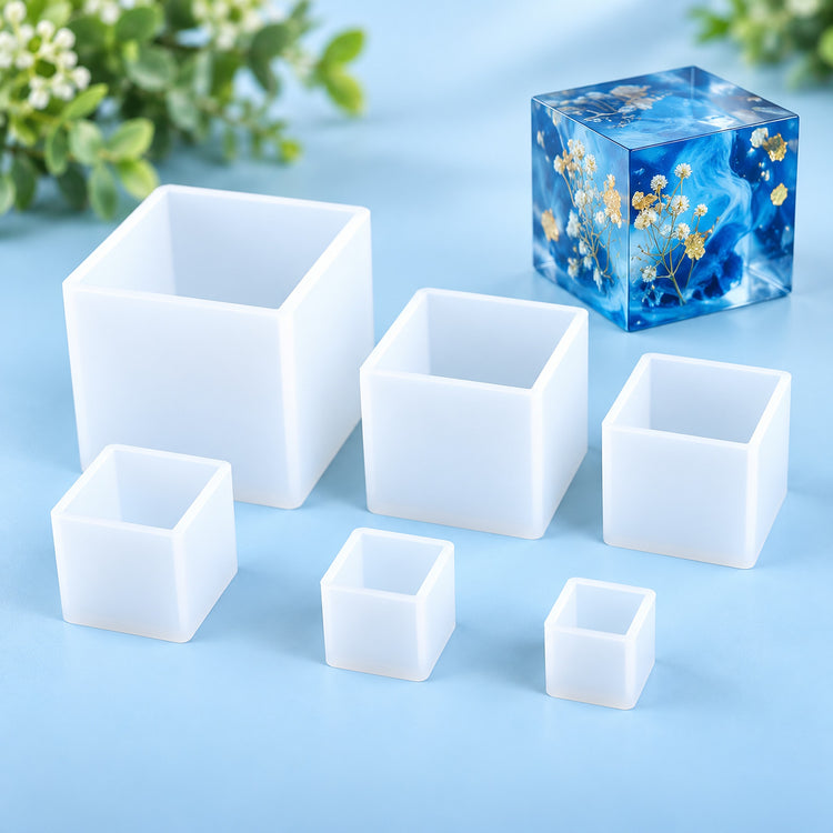 Six translucent white silicone mold boxes in different sizes arranged on a light blue surface, with a blue resin cube containing small yellow flowers placed behind them.