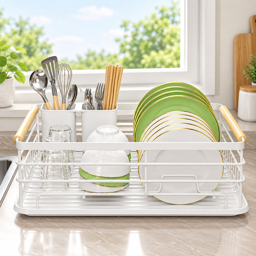 A white dish drying rack with gold-toned handles sits on a kitchen counter in front of a window. The rack holds white plates with gold trim, green plates, bowls, and utensils in holders.