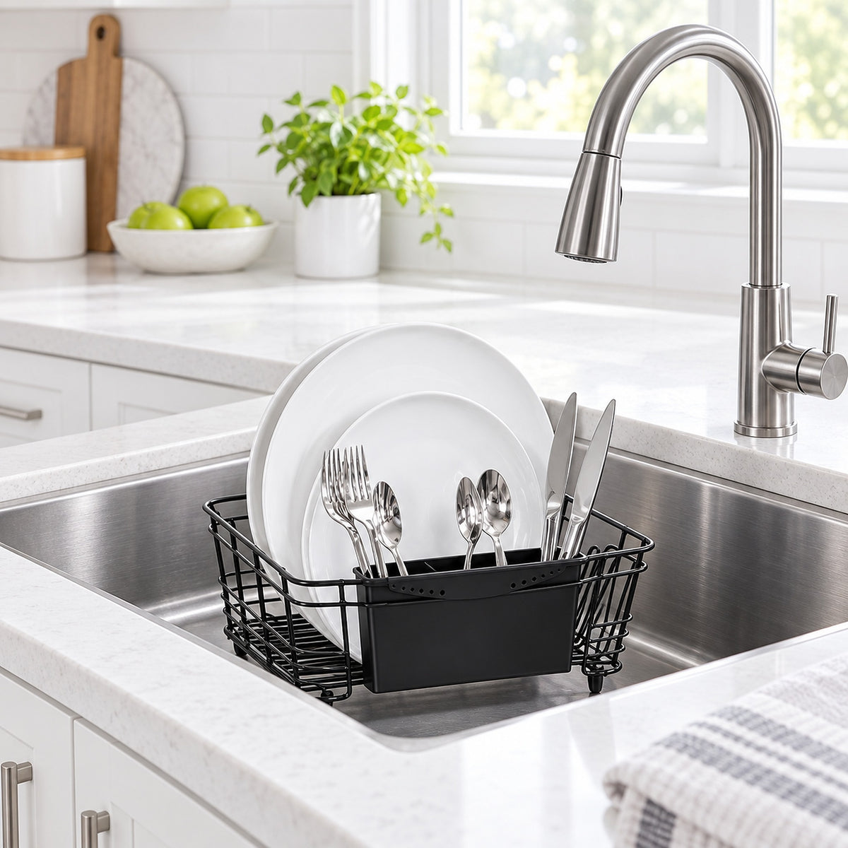 A black wire dish rack with a black storage bin holding white plates and silver cutlery, placed in a stainless steel kitchen sink with a modern faucet. The sink is set in a bright, clean kitchen with white cabinets, a tiled backsplash, and a window in the background.