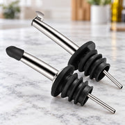 Two stainless steel oil pourers with black rubber stoppers resting on a white marble countertop in a bright kitchen setting.